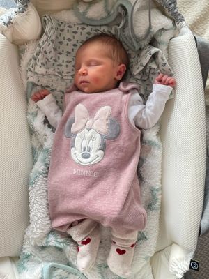 Ein Babymädchen schläft friedlich in einem Kinderbett und trägt ein rosa Minnie-Mouse-Kleidchen mit einer Schleife auf der Brust. Das Bett ist mit einer weichen Decke bedeckt.