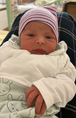 Ein Neugeborenes ist in einem weißen Outfit und einer pink-weiß gestreiften Mütze gekleidet. Das Baby schaut mit leicht geöffnetem Mund geradeaus.