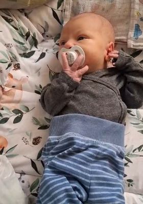 Ein Baby liegt auf einem Bett mit einer Blumendecke, trägt ein graues Langarmshirt und blaue gestreifte Hosen und hält einen Schnuller in der rechten Hand.