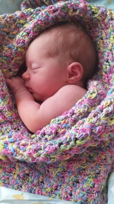 Ein Baby schläft friedlich auf einer bunten gestrickten Decke. Das Gesicht des Babys ist zur Seite gedreht und eine Hand ruht auf der Decke. Die Decke besteht aus verschiedenen Farben, darunter Pink, Blau, Gelb und Grün.