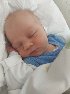 Ein schlafendes Neugeborenes in einem Krankenhausbett, das ein blaues Shirt und ein weißes Langarm-Body trägt. Das Baby hat ein Krankenhausarmband am Handgelenk.