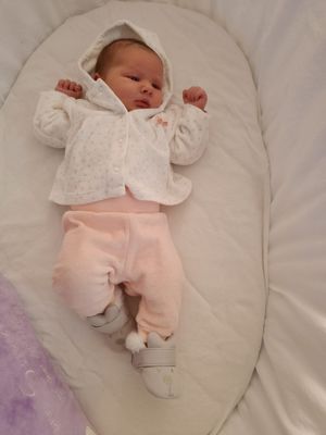 Ein Babymädchen liegt auf einer weißen Decke und trägt ein rosa Outfit mit einer weißen Jacke und grauen Stiefeln mit weißen Quasten.