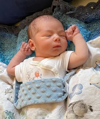 Ein Neugeborenes schläft friedlich auf einer blau-weißen Decke mit einem blauen Tuch um den Hals. Das Baby trägt ein weißes Shirt mit einem Ballon-Design.