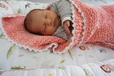 Ein Neugeborenes schläft friedlich auf einer rosa-weißen gehäkelten Decke mit einem grauen Shirt. Die Hand des Babys ist leicht erhoben.