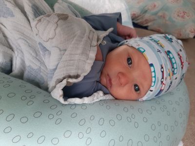Ein Babyjunge liegt auf einer blauen Decke mit einem Punktemuster, trägt ein blaues T-Shirt und eine Mütze mit LKW-Design. Er ist in ein weißes Tuch gewickelt und schaut direkt in die Kamera.