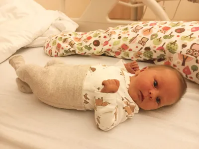 Ein Baby liegt auf einem Krankenhausbett und trägt ein weißes Strampler mit Tierdruck. Ein bunter Schlafsack befindet sich hinter dem Baby.