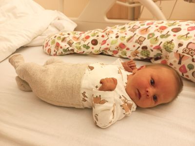 Ein Baby liegt auf einem Krankenhausbett und trägt ein weißes Strampler mit Tierdruck. Ein bunter Schlafsack befindet sich hinter dem Baby.