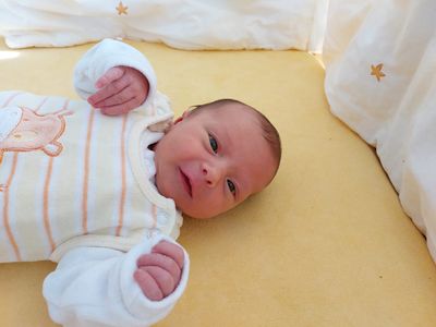 Ein lächelndes Neugeborenes liegt auf einer gelben Decke und trägt ein weiß-orange gestreiftes Strampler. Die Hände des Babys sind leicht gekrümmt und es schaut direkt in die Kamera.