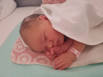 Ein Neugeborenes schläft friedlich in einem Krankenhausbett, bedeckt mit einer weißen Decke und trägt ein rosa Armband. Die Hand des Babys ruht auf seiner Wange.