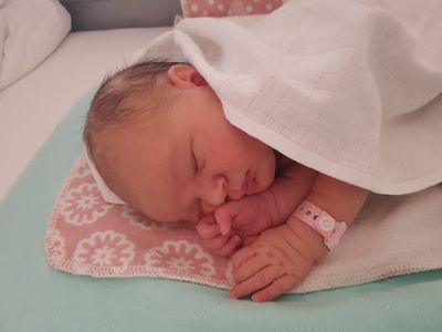 Ein Neugeborenes schläft friedlich in einem Krankenhausbett, bedeckt mit einer weißen Decke und trägt ein rosa Armband. Die Hand des Babys ruht auf seiner Wange.