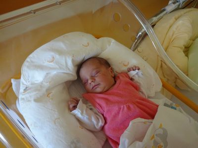 Ein Babymädchen schläft friedlich in einem Krankenhausbettchen, trägt ein rosa Kleid und ist von weißen Kissen umgeben.