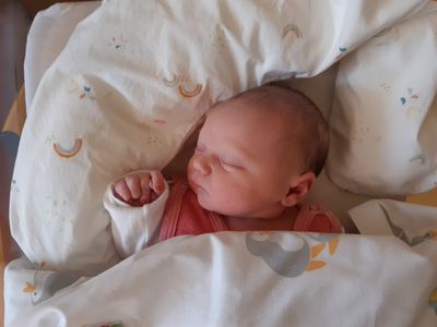 Ein Neugeborenes schläft friedlich auf einem Bett, bedeckt mit einer weißen Decke mit bunten Mustern. Das Gesicht des Babys ist leicht zur Seite gedreht, und eine Hand ruht sanft auf der Decke.