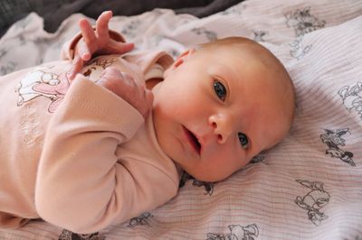 Ein Baby liegt auf einem Bett mit einer Blumendecke, trägt ein rosa Langarmshirt und schaut direkt in die Kamera mit einem Lächeln.
