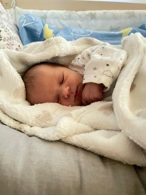 Ein Neugeborenes schläft friedlich auf einem Bett, bedeckt mit einer weißen Decke. Das Baby trägt ein weißes Outfit mit Sternen und Wolken.