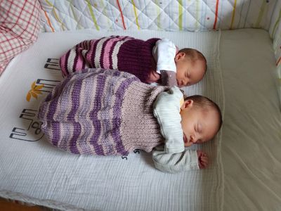 Zwei Babys schlafen auf einem Bett, eines trägt ein lila-weißes gestricktes Outfit, das andere ein weiß-grau gestreiftes Hemd.