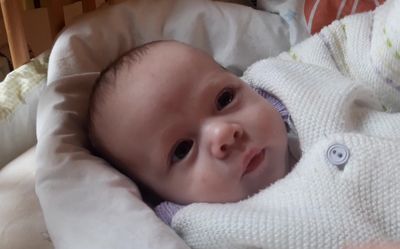 Ein Nahaufnahme des Gesichts eines Babys, in eine weiße Decke gewickelt, auf einem weißen Kissen liegend. Das Baby hat dunkle Haare und schaut direkt in die Kamera.