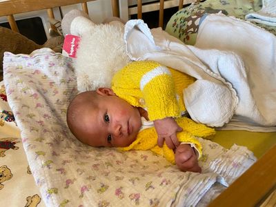 Ein Baby liegt auf einer blumengemusterten Decke und trägt einen gelben Pullover mit weißen Streifen. Ein weißes Plüschtier ist in der Nähe.