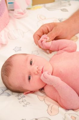 Ein Neugeborenes liegt auf einem Bett, während eine Person seine Hand hält. Das Baby schaut mit neugierigem Ausdruck zur Kamera.