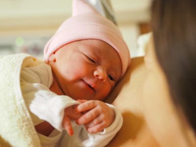 Ein Neugeborenes mit rosa Mütze wird von einer Frau gehalten, beide schauen in die Kamera.
