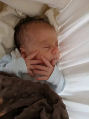 Ein Neugeborenes schläft friedlich, eingewickelt in eine weiße Decke und einen braunen, strukturierten Stoff. Die Hände des Babys liegen sanft über seinem Gesicht.
