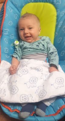 Ein lächelndes Baby in einer blau-weißen Decke mit Löwenmuster, das ein grünes Langarmshirt trägt, an dessen Kragen ein Schnuller befestigt ist.