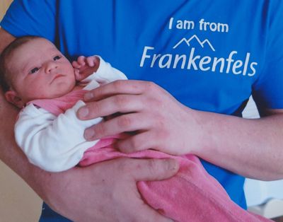 Eine Person hält ein Neugeborenes in den Armen, das Baby ist in eine rosa Decke gewickelt. Die Person trägt ein blaues Shirt mit den Worten 'Ich komme aus Frankenfels'.