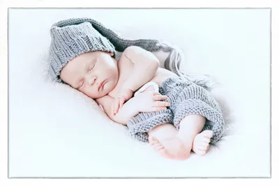 Ein schlafendes Baby in einer gestrickten Mütze und einem Strampler, auf einer weißen Decke liegend. Die Augen des Babys sind geschlossen, und es scheint in einem friedlichen Schlaf zu sein.