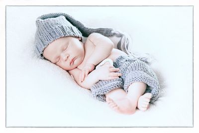 Ein schlafendes Baby in einer gestrickten Mütze und einem Strampler, auf einer weißen Decke liegend. Die Augen des Babys sind geschlossen, und es scheint in einem friedlichen Schlaf zu sein.
