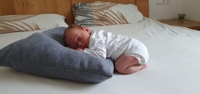 Ein Neugeborenes schläft auf einem blauen Kissen auf einem Bett, trägt ein weißes Outfit und eine Windel.