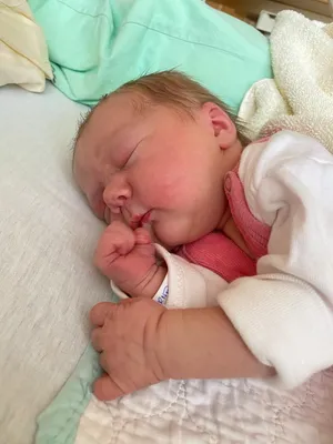 Ein Neugeborenes schläft friedlich auf einem Bett, trägt ein rosa Hemd und eine weiße Decke. Das Baby hat einen Schnuller im Mund und hält die Hand vors Gesicht.