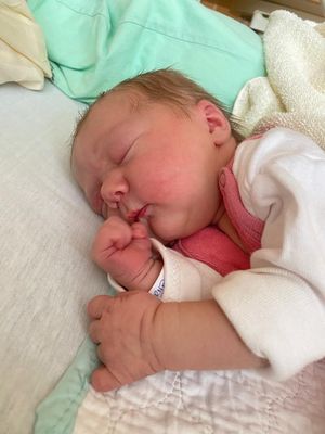 Ein Neugeborenes schläft friedlich auf einem Bett, trägt ein rosa Hemd und eine weiße Decke. Das Baby hat einen Schnuller im Mund und hält die Hand vors Gesicht.