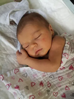 Ein Neugeborenes schläft friedlich auf einer weißen Decke mit roten Herzdesigns. Das Baby ist in einen weißen Stoff gewickelt und hat einen kleinen Verband am rechten Arm.