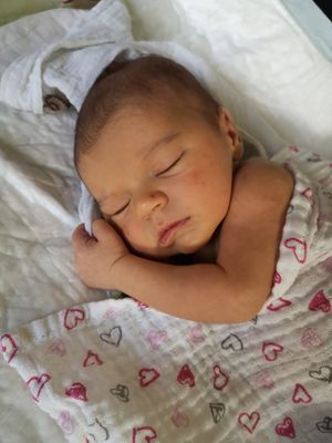 Ein Neugeborenes schläft friedlich auf einer weißen Decke mit roten Herzdesigns. Das Baby ist in einen weißen Stoff gewickelt und hat einen kleinen Verband am rechten Arm.