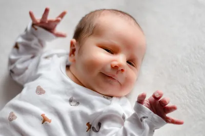 Ein lächelndes Baby liegt auf einer weißen Oberfläche und trägt ein weißes Onesie mit Tierdruck. Die Hände des Babys sind erhoben, und es scheint etwas mit Interesse zu betrachten.