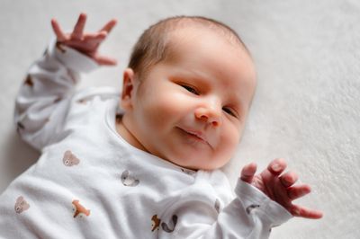 Ein lächelndes Baby liegt auf einer weißen Oberfläche und trägt ein weißes Onesie mit Tierdruck. Die Hände des Babys sind erhoben, und es scheint etwas mit Interesse zu betrachten.