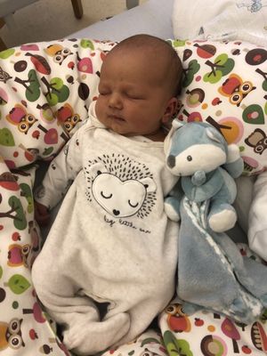 Ein Baby schläft in einem Kinderbett mit einer bunten Decke und einem blauen Plüschtier. Das Baby trägt einen weißen Strampler mit Herzdesign.