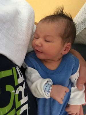 Ein Neugeborenes, das ein blaues Shirt und weiße lange Ärmel trägt, wird von einer Person gehalten, wahrscheinlich ein Elternteil. Das Baby hat eine medizinische Armband am Handgelenk.