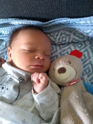 Ein schlafendes Baby mit einem Teddybären, beide liegen auf einer blauen Decke mit geometrischem Muster.