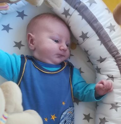 Ein Baby liegt auf einem Bett mit einem blauen Shirt mit Weltraumdesign, umgeben von sterngemusterten Kissen.