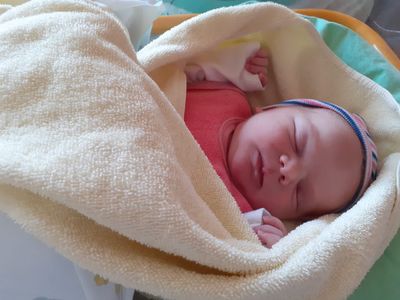 Ein Neugeborenes schläft friedlich in einer weißen Decke eingewickelt, trägt ein rosa Hemd und eine blau-weiße Kopfband.