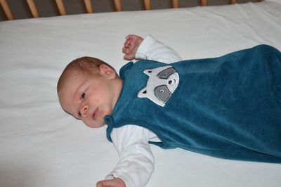 Ein Baby liegt auf einem weißen Laken und trägt ein blaues Overall mit einem Waschbär-Design. Das Baby hat ein leichtes Lächeln und schaut direkt in die Kamera.