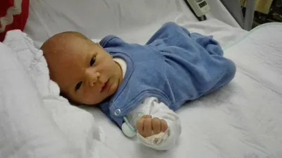 Ein Neugeborenes liegt auf einem Krankenhausbett, trägt ein blaues Outfit und hält einen Schnuller in der Hand.