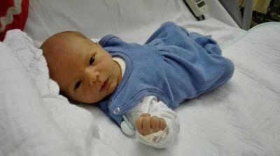 Ein Neugeborenes liegt auf einem Krankenhausbett, trägt ein blaues Outfit und hält einen Schnuller in der Hand.