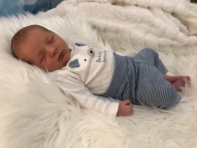 Ein Neugeborenes schläft friedlich auf einer weißen Pelzdecke und trägt ein weiß-blau gestreiftes Outfit mit einem Bärendesign.