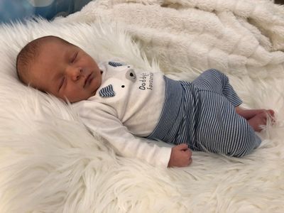 Ein Neugeborenes schläft friedlich auf einer weißen Pelzdecke und trägt ein weiß-blau gestreiftes Outfit mit einem Bärendesign.