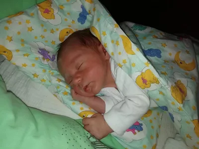 Ein Neugeborenes schläft friedlich unter einer Decke mit Zeichentrickfiguren und Sternen. Das Baby trägt ein weißes Langarmshirt.