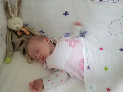 Ein Babymädchen schläft auf einem Bett mit einer weißen Decke und einem Plüschtier. Sie trägt ein rosa Outfit mit Schmetterlingsdesign.