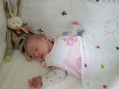 Ein Babymädchen schläft auf einem Bett mit einer weißen Decke und einem Plüschtier. Sie trägt ein rosa Outfit mit Schmetterlingsdesign.
