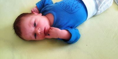 Ein Baby liegt auf einer gelben Oberfläche und trägt ein blaues Shirt mit einer handgezeichneten Zeichnung. Das Baby hat einen neugierigen Ausdruck und berührt sein Gesicht.