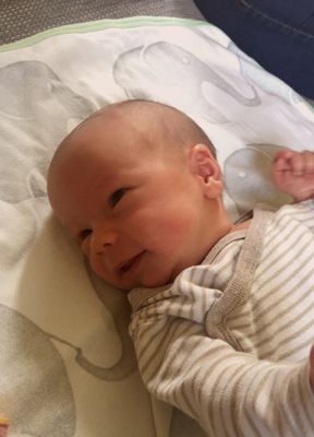 Ein lächelndes Baby mit kahlem Kopf liegt auf einem Bett mit gemusterter Decke. Das Baby trägt einen gestreiften Strampler.